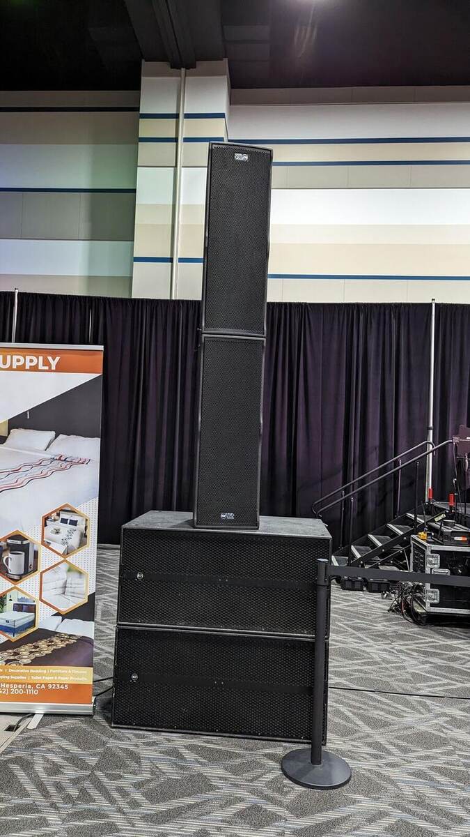 A tall vertical line-array speaker stack on a subwoofer at a trade show/convention hall, beside a 'SUPPLY' pro-audio banner and black drape