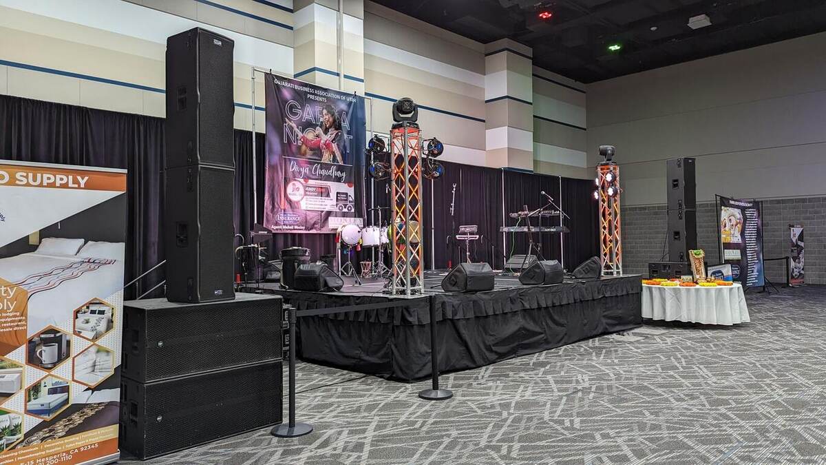 A trade-show booth with a small riser stage, drum kit, orange truss, line-array speakers, monitors, and a pro-audio 'SUPPLY' banner in a convention center