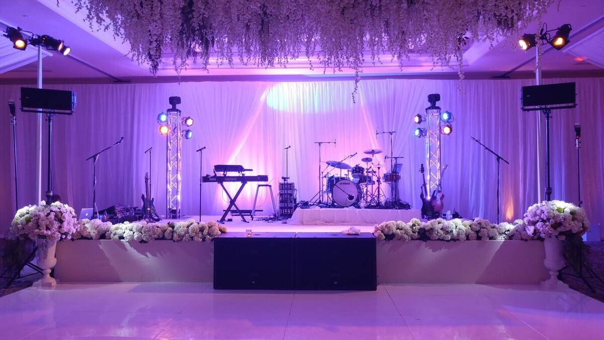 Indoor event stage set for a live band (keyboard, drum kit, guitars) with purple/pink uplighting, white draped backdrop, hanging floral greenery, and flower arrangements lining the stage front