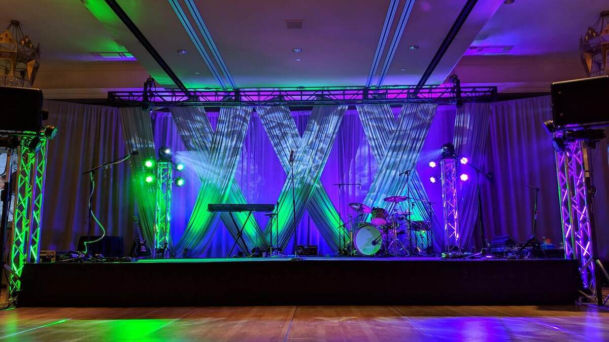 Empty indoor stage with green and purple lighting, crisscrossed white draping backdrop, truss towers, keyboard and drum kit, on a wood dance floor