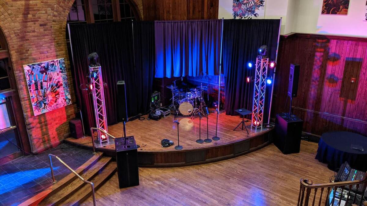 Empty band stage in a brick-walled hall with dark backdrop, blue/red uplighting, truss towers, drum kit, and multiple mic stands, viewed from a balcony
