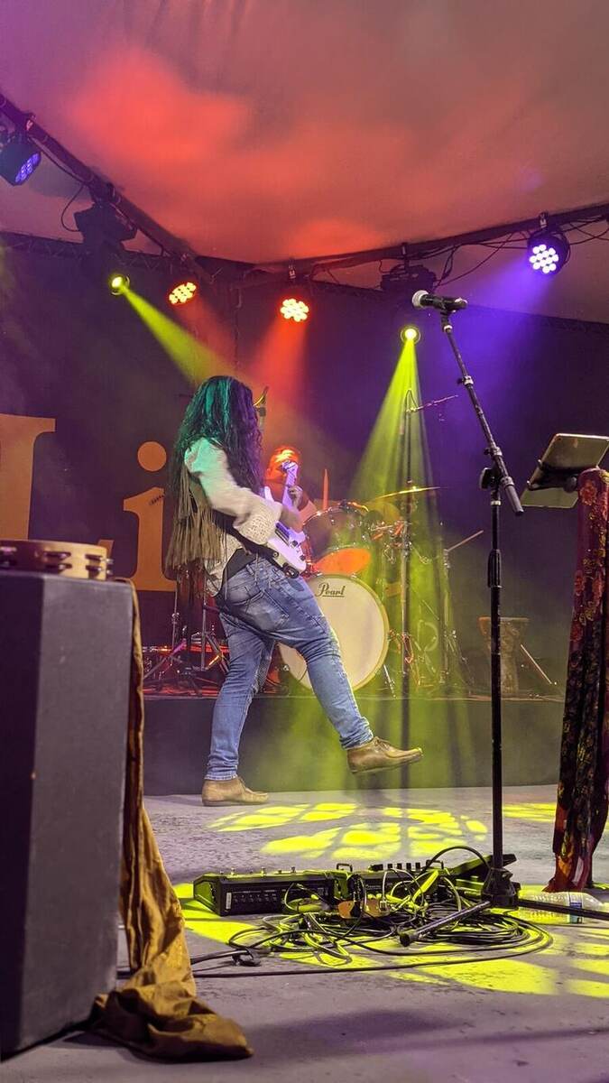 A long-haired guitarist mid-performance on stage lit with yellow, purple, and orange beams; a drum kit, monitor wedge, and cables with a mixer sit in the foreground