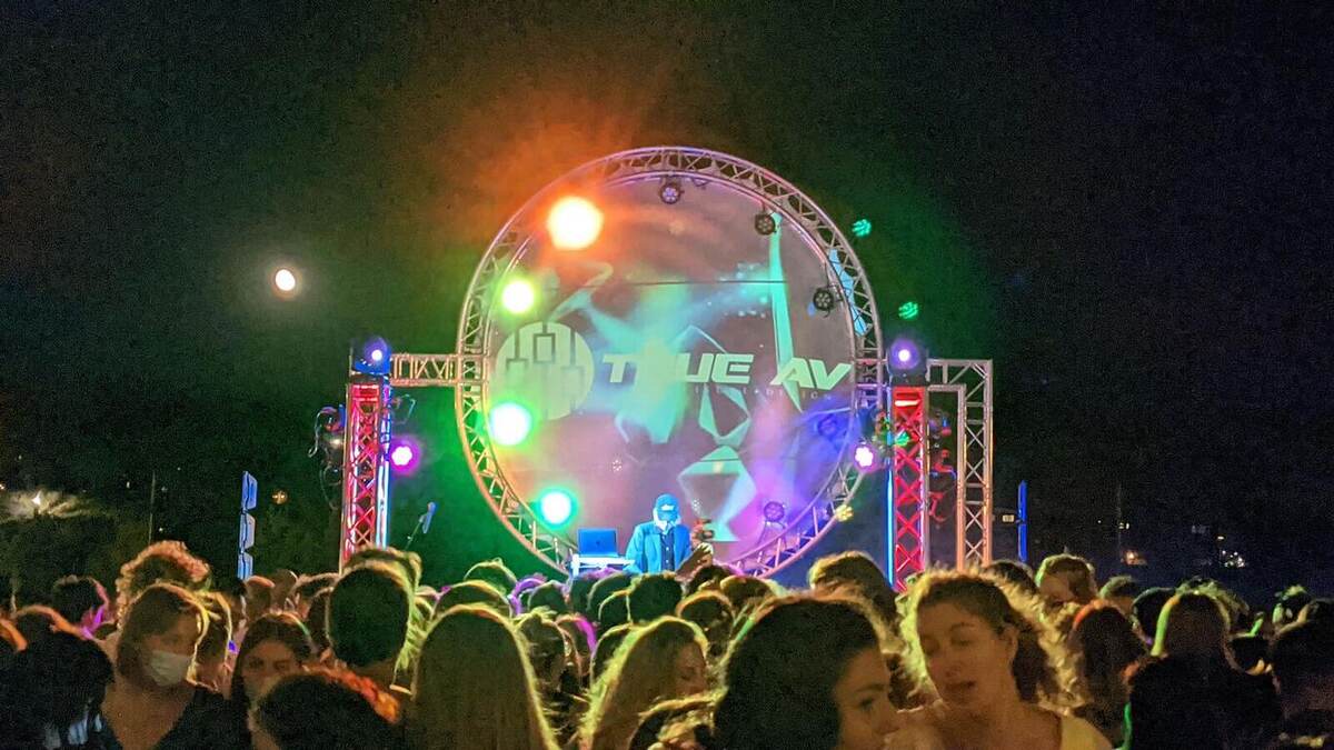 A nighttime outdoor crowd faces a brightly lit circular-truss stage displaying the 'True AV' logo amid colorful lighting
