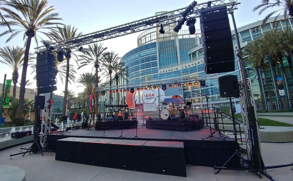 Outdoor plaza stage in front of the Anaheim Convention Center with truss rigging, line arrays, band gear, palm trees, and a branded 'ARA' backdrop sign