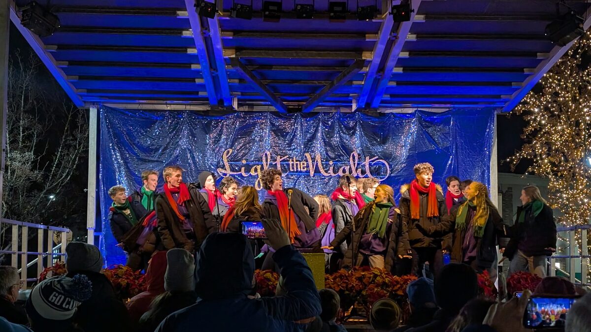 Carolers in red and green scarves performing under blue stage lighting at a Light the Night holiday event