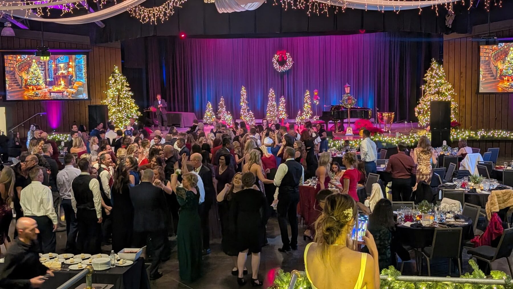 A packed corporate holiday gala, guests dancing in formal attire before a stage lined with lit Christmas trees and LED screens
