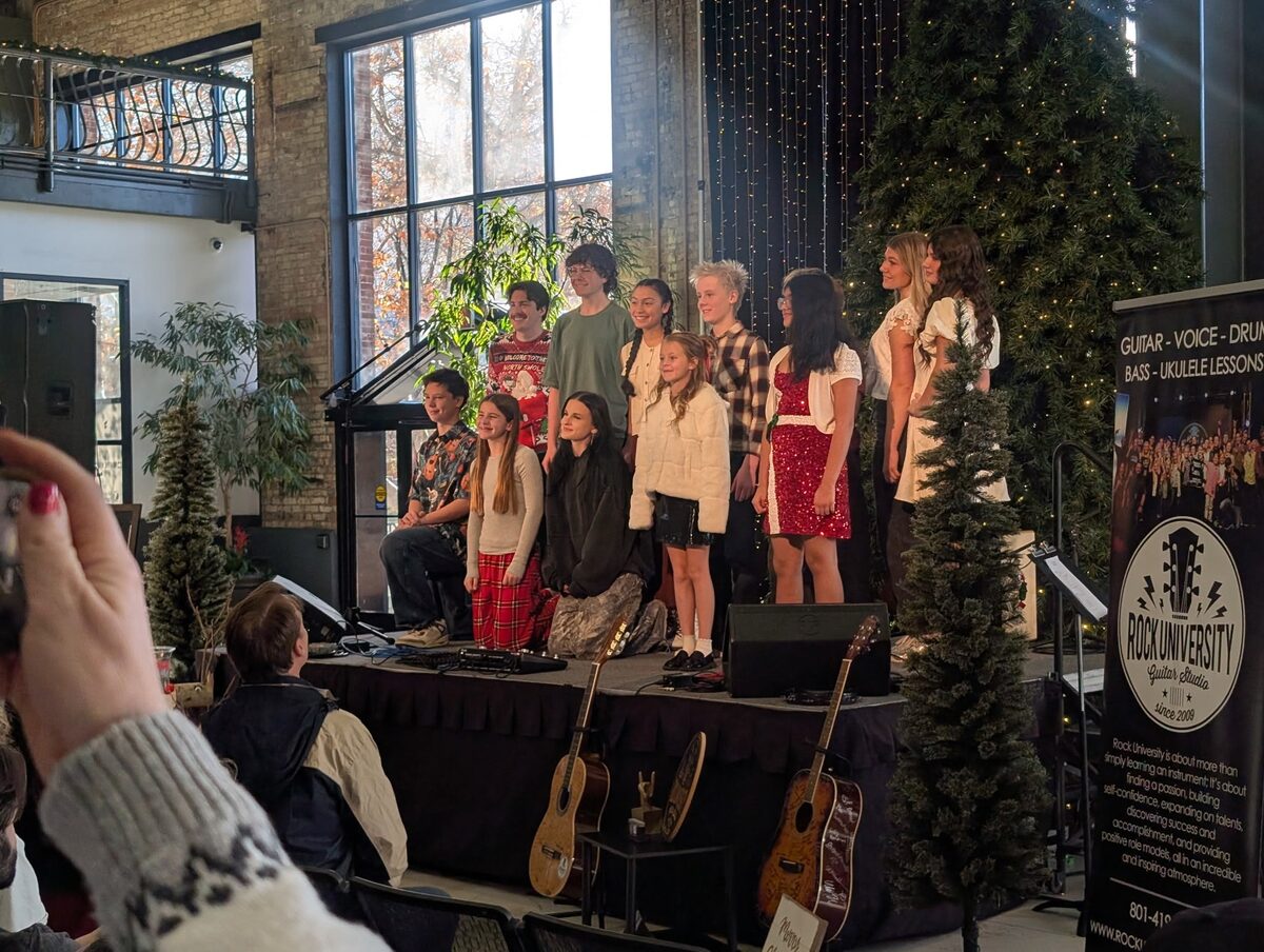 A Rock University holiday showcase, young performers on a string-lit stage beside a tall Christmas tree