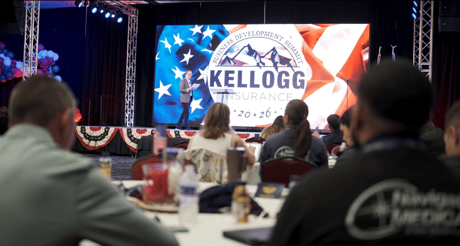 A keynote speaker in front of the branded Kellogg Insurance LED wall with live IMAG