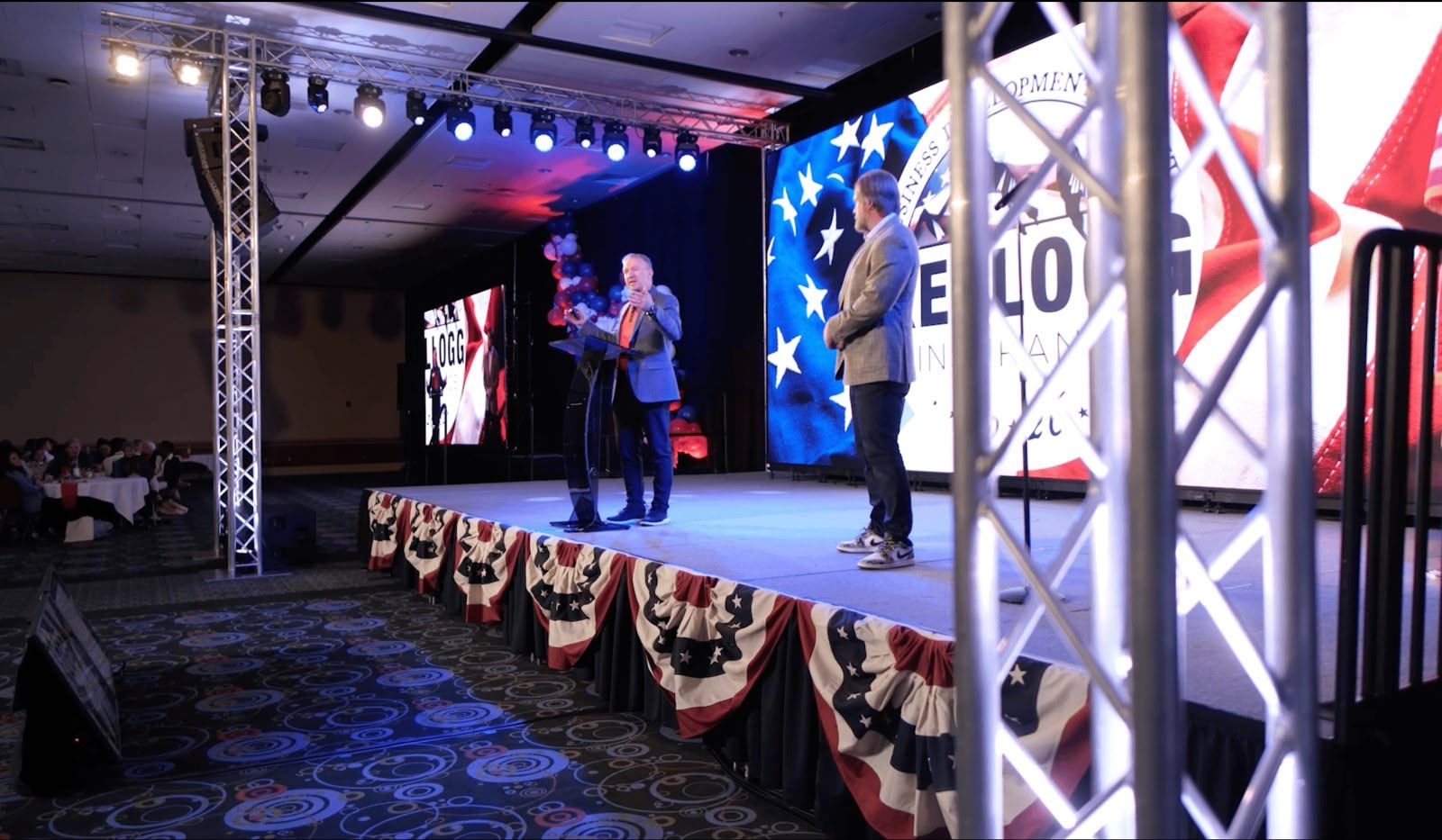 A branded corporate keynote on a patriotic LED-lit stage, a speaker at a clear podium with every word heard