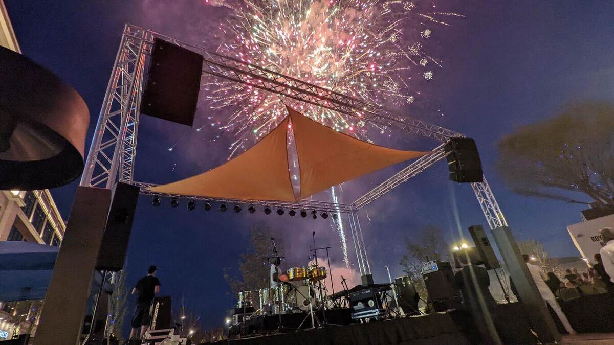 Fireworks burst above a truss-framed outdoor stage with a sail shade and drum kit while a lone figure watches; dramatic night sky