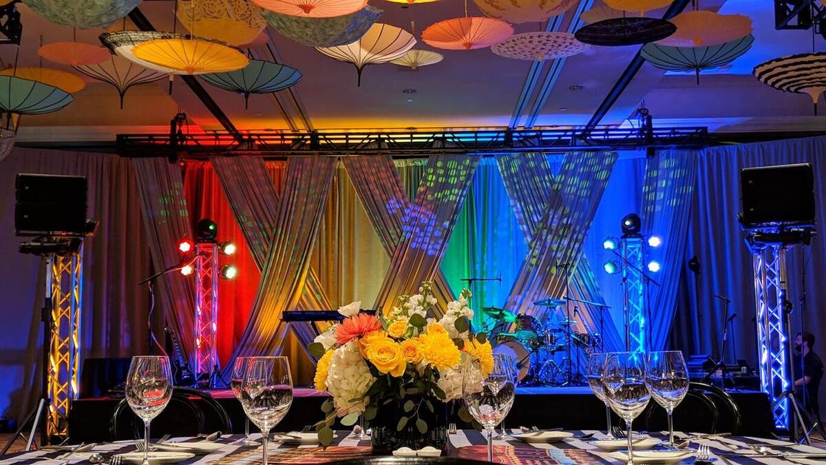 Ballroom head-table view with a rainbow-lit stage backdrop (red, green, blue washes), band gear, truss towers, colorful parasols on the ceiling, and a floral centerpiece with wine glasses in front