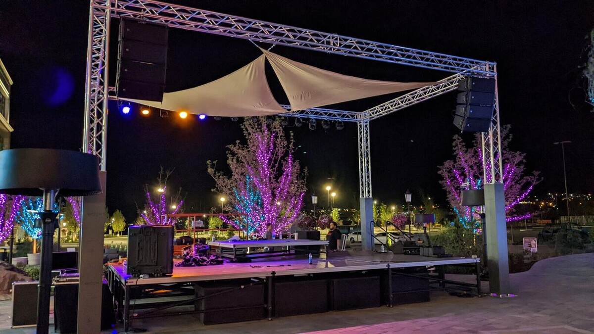 A compact outdoor platform stage under a truss goalpost with a fabric sail shade and stage lighting, set against a plaza with purple/pink-lit trees at night