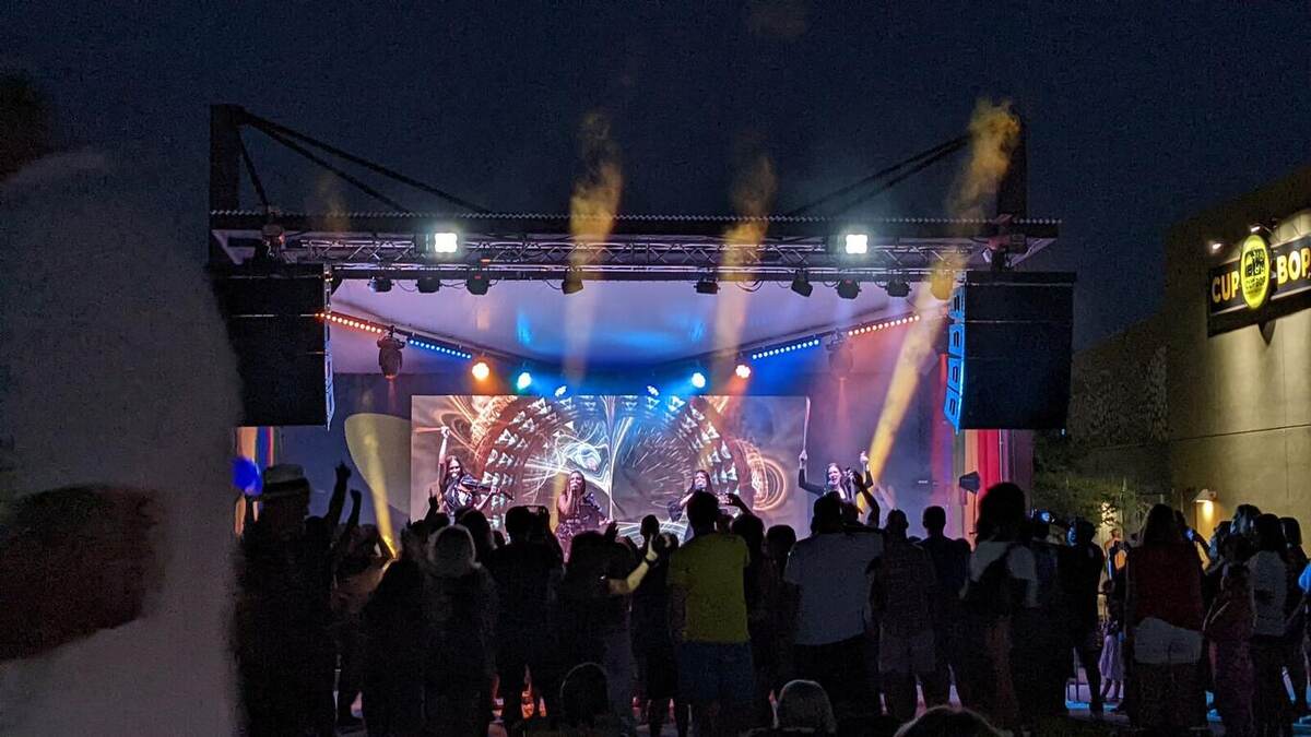 A nighttime outdoor concert with a band on stage, colorful lighting, atmospheric fog jets, and an animated LED video wall, with an enthusiastic crowd raising hands in front of a shopping-center building