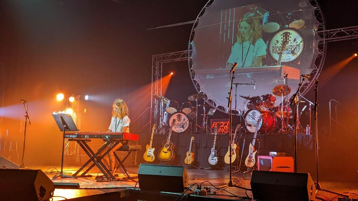 A keyboardist performs on a stage lit in warm orange, with a full drum kit, guitar rack, and a large circular video screen showing a live close-up of a band member