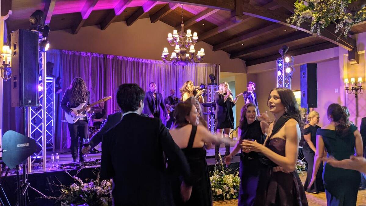 Formal-dressed guests dancing in front of a live band under purple lighting and a chandelier in a rustic-beamed ballroom, with truss uplights framing the stage