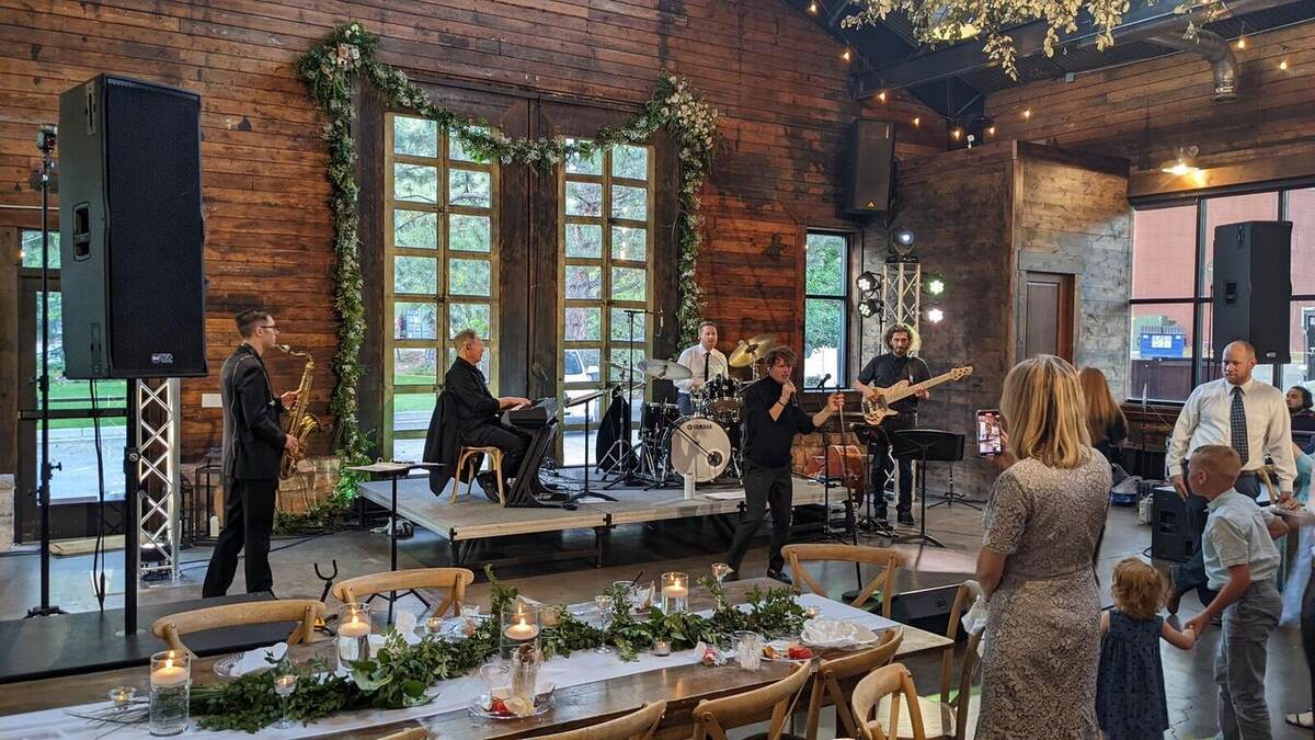A jazz/horn band performs on a low riser stage inside a rustic wood-beam venue with greenery-draped windows and candlelit banquet tables in the foreground